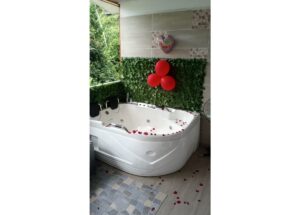 Romantic outdoor bathroom scene with a white corner jacuzzi tub, red rose petals scattered on the tub and floor, and red balloons tied to a green leafy wall beside a heart-shaped sign reading 'Te amo'