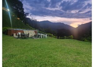 Outdoor patio with a raised hot tub and stone wall, plastic chairs and tables on a green lawn at sunset overlooking hills.
