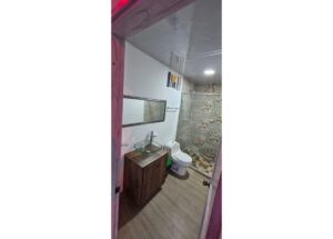 Small bathroom with a wooden vanity and glass vessel sink, mirror above, and a tiled shower stall to the right; toilet beside the vanity.