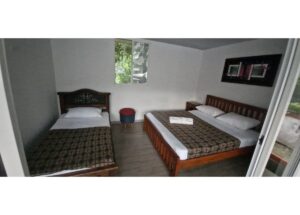 Hotel room with a single bed on the left and a double bed on the right, wood headboards, patterned bedcovers, a small round stool between, and a window with wall art above the double bed