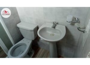 Small bathroom with a toilet beside a pedestal sink, tiled walls, and a wall soap holder (logo in the top-left).