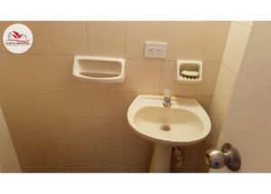 Small bathroom sink area with pedestal sink, wall soap dish containing a bar of soap, and an electrical outlet on tiled wall; watermark logo in top-left corner.