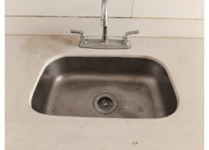 Stainless steel kitchen sink with a dual-handle faucet; dirty countertop and grime around the basin.