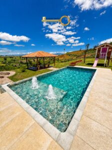 Rectangular pool with two fountains, bordered by a stone deck, with a thatched gazebo and a pink-slide playhouse on a sunny hillside.