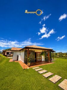 Modern single-story house with a stone facade, terracotta roof, and a tiled patio surrounded by a green lawn under a bright blue sky; logo watermark reads Riveros Ortiz Real Estate.