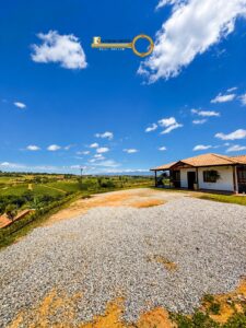 Rural property with a white house and orange-tiled roof on the right, gravel driveway in foreground, rolling green fields, and a bright blue sky with clouds; logo for Riveros Ortiz Real Estate appears in the sky above the scene.
