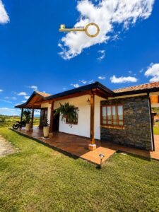 Single-story house with a covered veranda, terracotta tiles, stone accent wall, potted plants, and a motorcycle under the front porch against a blue sky with clouds (real estate branding in the sky).