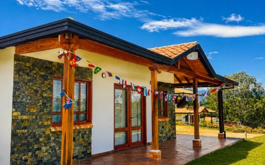 Sunny single‑story house with a stone façade, wooden columns, and a tiled porch decorated with international flags.