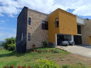 Modern two-story house with stone facade, a mustard-yellow boxy wing, and a two-car carport with SUVs parked inside.
