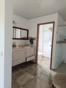 Bathroom with a double-sink vanity, large wooden-framed mirror, and marble floor tiles; doorway reveals toilet and a glass-walled shower area.