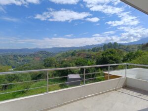 Balcony railing overlooking a lush green hillside valley under a blue sky with scattered clouds.