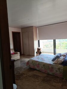 Bedroom with floral quilted bed, a tall wooden wardrobe, and a beige chair with a teddy bear by the window looking out onto a balcony.