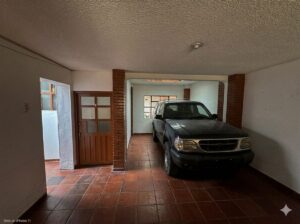 Garage interior with a dark blue SUV parked on red tile floor between brick columns and a wooden door on the left side of the image.