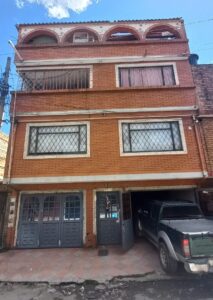 Brick four-story building with arched rooftop openings and barred windows; a pickup truck parked under a carport on the right and a tiled sidewalk in front.