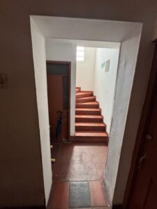 Indoor stairwell with red steps winding up beside a white wall and door, exit sign visible on the wall ahead.