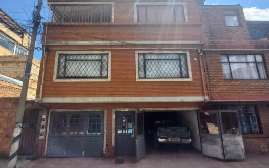 Three-story red brick building with a ground-floor carport; a car is parked inside the driveway beneath the building on a sunny day.