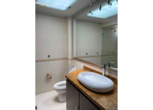 Modern bathroom with an oval vessel sink on a brown marble countertop, large mirror, and beige tiled walls. A toilet sits beside the vanity. A shower is visible in the reflection above.