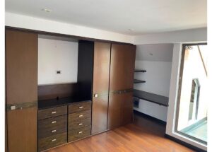 Built-in wooden wardrobe with multiple drawers and a small open shelf area, plus a side desk alcove in a room with wood flooring.