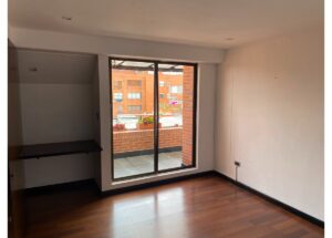 Empty room with hardwood floor, a built-in desk alcove on the left, and a sliding glass door opening to a brick balcony with potted plants; city buildings visible outside.