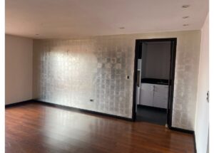 Empty living room with a silver tiled accent wall, wooden floor, and an open doorway to a small kitchen with white cabinets.