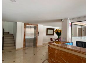Lobby reception with marble desk, stainless elevator doors framed by brown marble, and a left stairwell; sunflowers on a round table nearby.