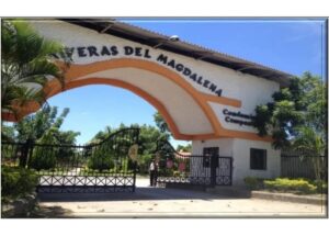 Gated entrance to a housing complex with a white building and orange curved arch, palm trees, and clear blue sky behind a metal gate, security booth nearby