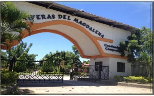 Gated entrance to a housing complex with a white building and orange curved arch, palm trees, and clear blue sky behind a metal gate, security booth nearby
