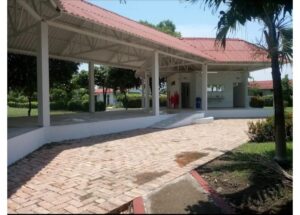 Open-air shelter with white columns and a red-tiled roof, brick-paved path, and manicured greenery nearby.