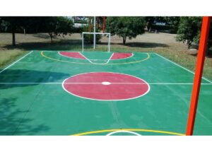 Outdoor basketball court with a green surface, red center circle and key shapes, white boundary lines, and a hoop frame in the background among trees.