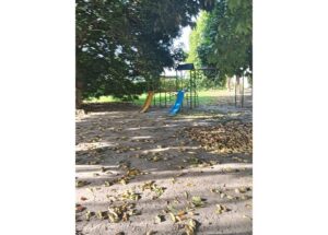 Playground with yellow and blue slides on a metal frame, shaded by trees; sandy ground scattered with fallen leaves.
