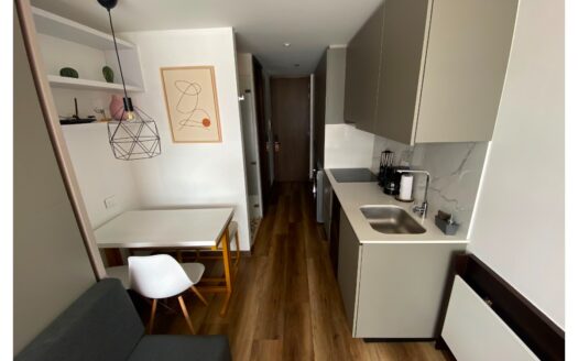 Open-plan kitchen with gray cabinets, white countertops, and a sink along a marble backsplash; a small dining table and white chair beside a gray sofa.