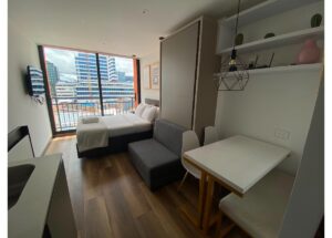 Bright studio apartment with a bed by a large window and balcony, cityscape view outside.